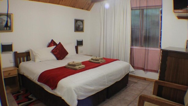 Een slaapkamer met een bed, nachtkastje, stoel, raam met gordijnen en een televisie.