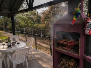 Een buitenbarbecue met een grill, witte plastic stoelen en een tafel met wijnglazen en een fles.