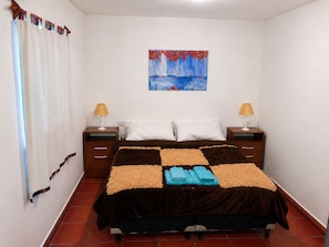 Una cama bien hecha con colcha a cuadros, dos lámparas de noche, un cuadro en la pared y una cortina en la ventana.