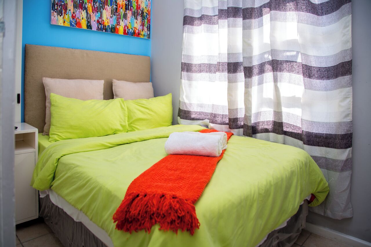 A neatly made bed with a green and orange blanket, a white pillow, and a striped curtain.