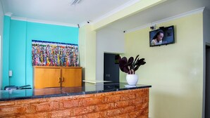 A reception area with a wooden counter, a colorful artwork on the wall, a television mounted on the wall, and a potted plant. - Room-relaxing Guest House in th Heart of (Cape Town)