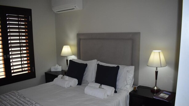 Una cama bien hecha con sábanas blancas, dos almohadas negras y un cabecero gris. Hay dos mesitas de noche con lámparas y una mesita auxiliar con una lámpara.