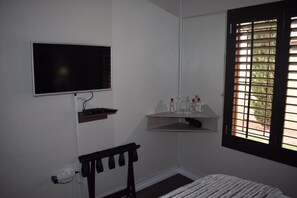 Una habitación con una cama, un televisor montado en la pared, una ventana con persianas y una estantería con botellas.