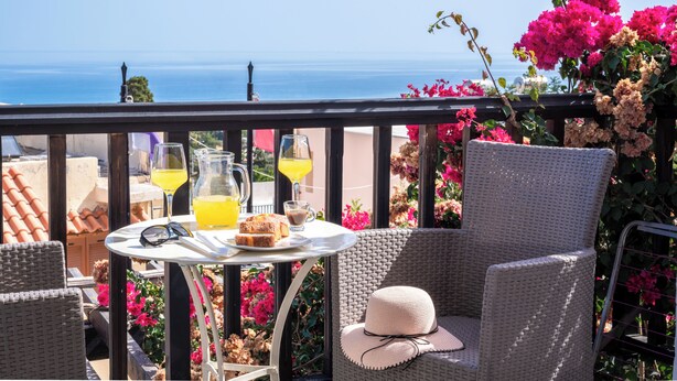 A balcony with wicker chairs, a table with drinks and food, and a view of the sea. - Korifi Suites Collection Apartment for Adults (Hersonissos)