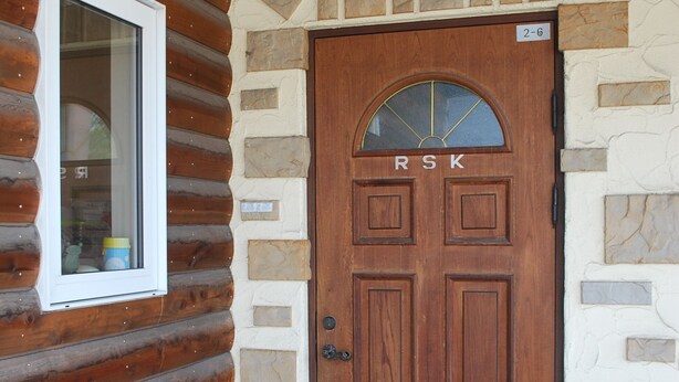 A wooden door with the letters "RSK" above it, a window to the left, and a stone wall with a number "2-6" sign.