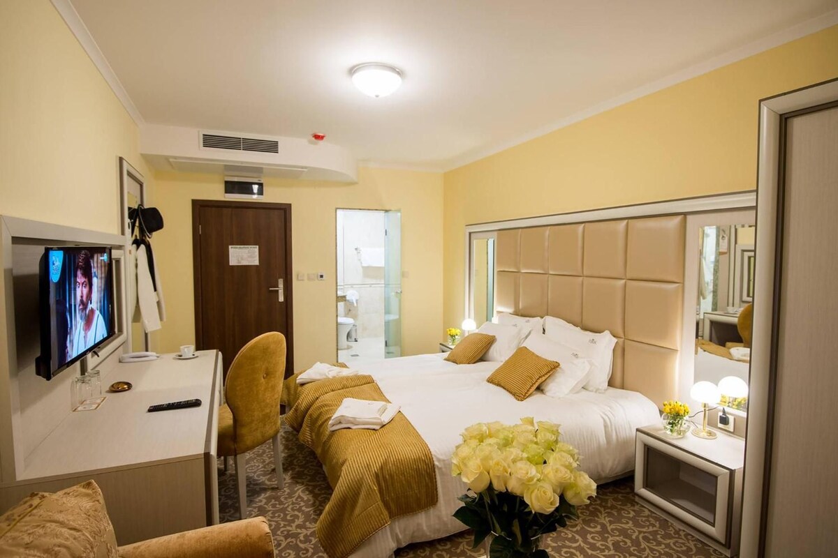 Quarto de hotel com cama, escrivaninha com televisão, cadeira, mesa de cabeceira com abajur e espelho.