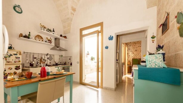 A kitchen with a wooden table, chairs, and a countertop with various items. There is a door leading to another room. - Little Home Holidays (Vernole)