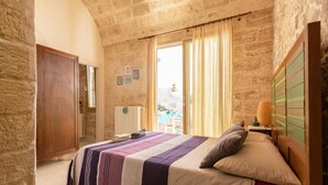 A bedroom with a stone wall, a bed with a striped bedspread, a wooden headboard, and a window with curtains. - Little Home Holidays (Vernole)