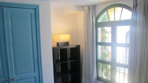A room with a blue door, a black shelving unit with a lamp, and a window with curtains. - Harbour 332 in Puerto De Mogan (Mogan)