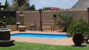 A swimming pool with a decorative fountain, surrounded by a fence and various plants. - Casa Villa Guest House Carletonville - Suite 2 (Merafong City)