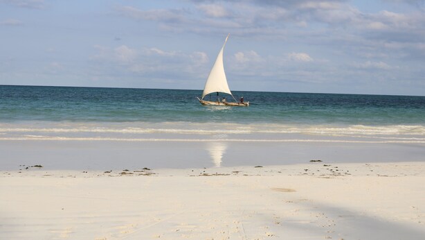 A boat with a white sail on a beach with clear water and a cloudy sky.