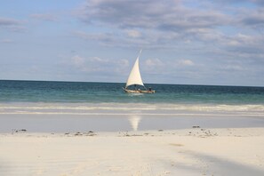 A boat with a white sail on a beach with clear water and a cloudy sky.