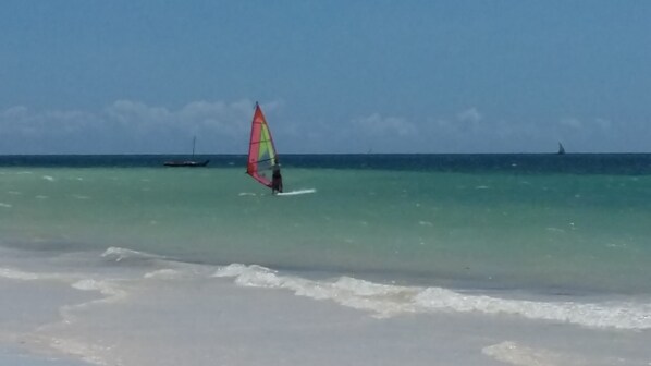 A beach with clear turquoise water, a person windsurfing, and a sailboat in the distance.