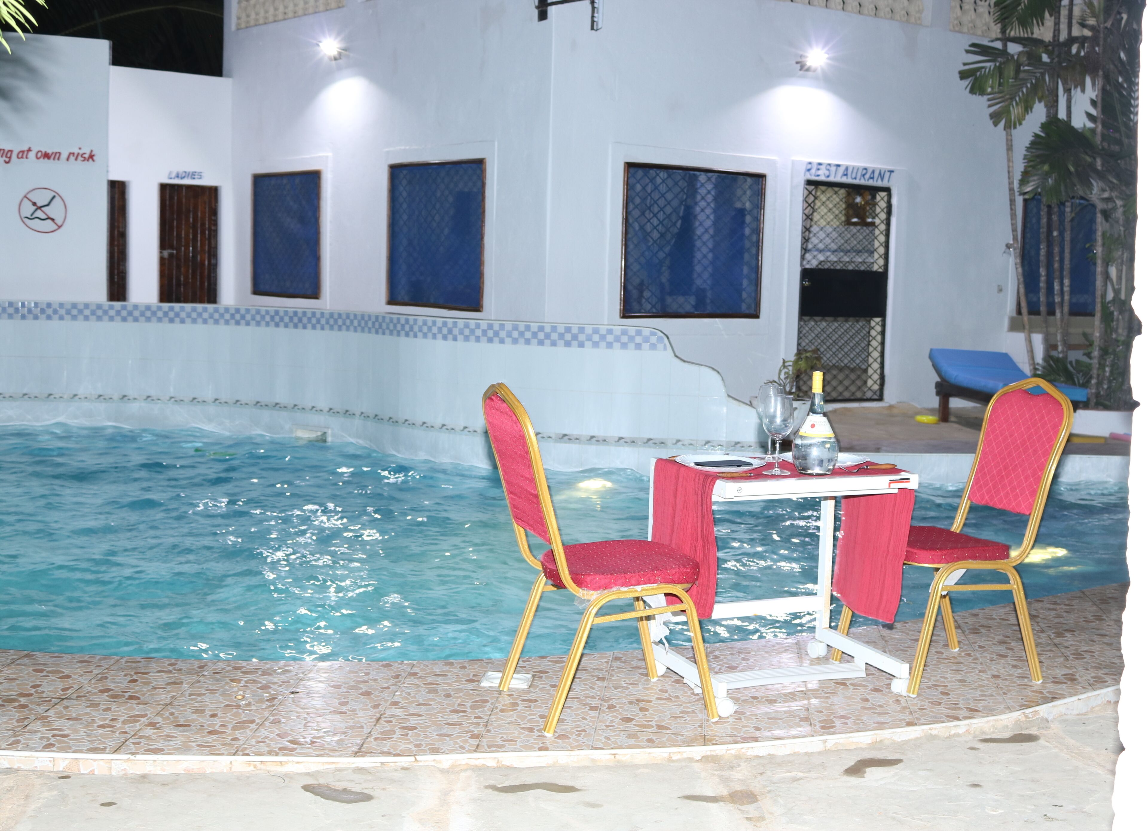 Un espace piscine intérieur avec deux chaises rouges et une table, un panneau indiquant la présence d’un restaurant, et une piscine à l’eau claire.