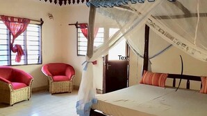 A room with a bed, two wicker chairs, a ceiling fan, and a window with curtains. - Room in Guest Room - A Wonderful Beach Property in Diani Beach Kenya - a Dream Holiday Place (Mombasa)
