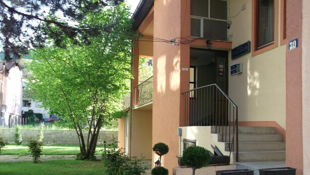 Ein Gebäude mit sandfarbener Fassade, einem Balkon mit Metallgeländer und einer Treppe, die zum Eingang führt. Vor dem Gebäude stehen Blumentöpfe.