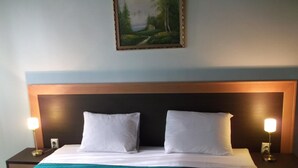 Ein ordentlich bezogenes Bett mit weißen Laken und einer petrolfarbenen Tagesdecke, flankiert von zwei Nachttischen mit Lampen, in einem Zimmer mit schlichtem Holz-Kopfteil und einem gerahmten Landschaftsbild an der Wand.