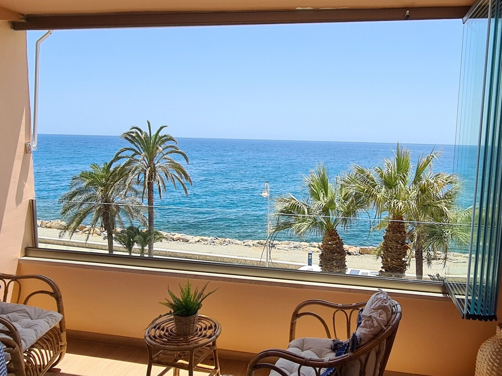 Un balcón con mobiliario de mimbre, una pequeña planta en maceta y vistas al mar y la playa.
