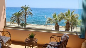Un balcón con mobiliario de mimbre, una pequeña planta en maceta y vistas al mar y la playa.