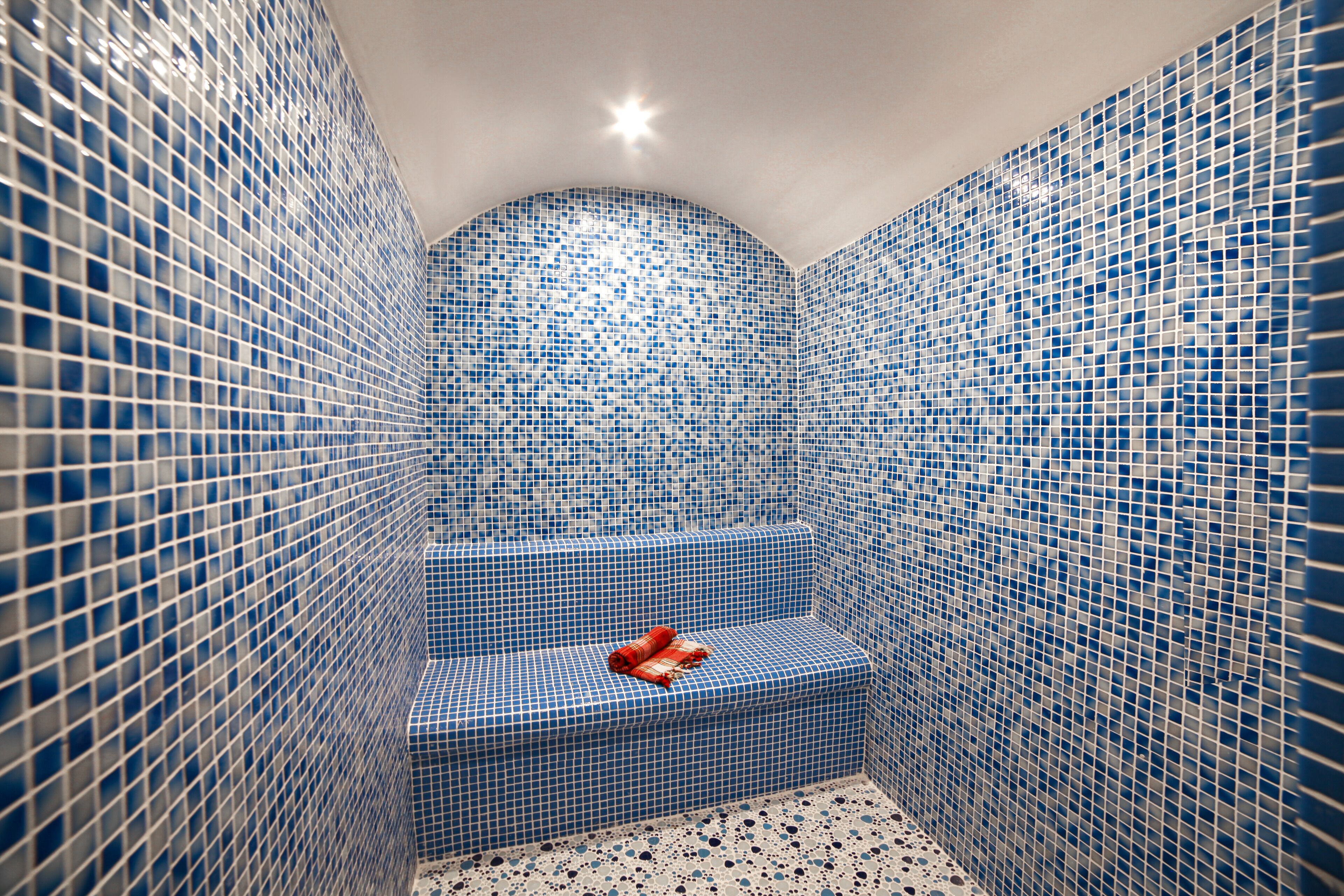 A small, enclosed space with blue mosaic tile walls and a tiled floor, featuring a single step with a red towel.