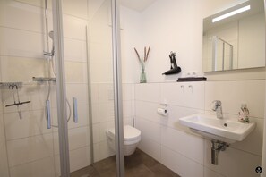 A modern bathroom with a glass shower enclosure, a white toilet, a sink with a mirror above it, and a small vase with dried plants on the wall. - Refugium 7 (Kellenhusen)