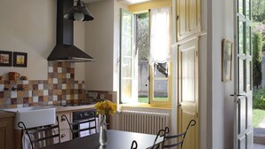 A kitchen with a stove, oven, and sink, a dining table with chairs, and a window with a view of greenery. - GITES GRAND CANISSOU (Carcassonne)
