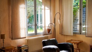 A cozy room with a large window, a radiator, a chair, a table, and a stack of books. - GITES GRAND CANISSOU (Carcassonne)