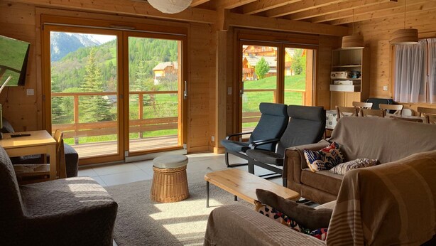 Una cabaña de madera acogedora con un amplio ventanal corredera que da a un balcón, una zona de estar confortable y un televisor sobre una mesa de madera.