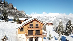 Una cabaña de madera con balcón, rodeada de árboles nevados y montañas al fondo.