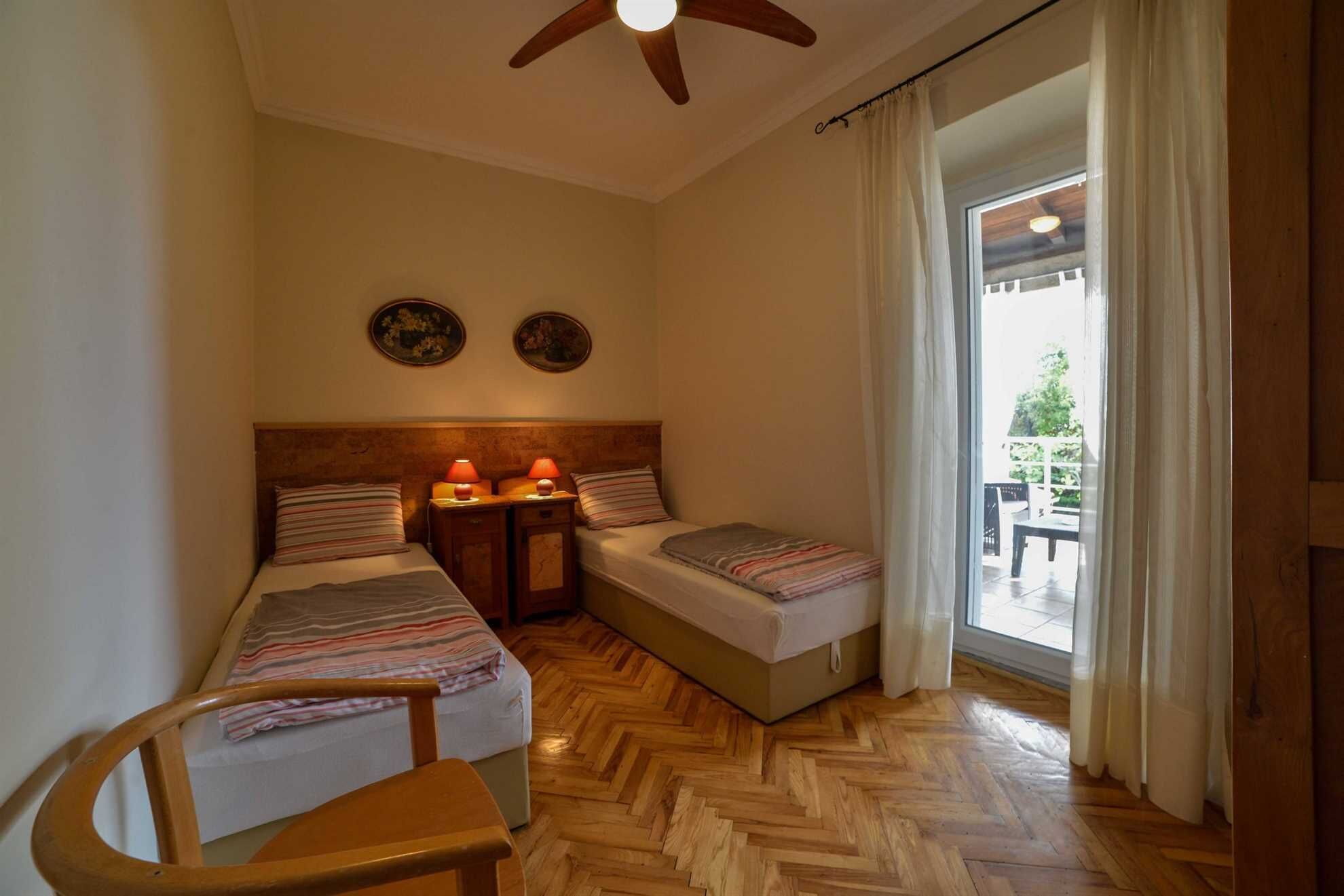 A bedroom with two single beds, a wooden chair, a ceiling fan, and a view of the outdoors through a window.