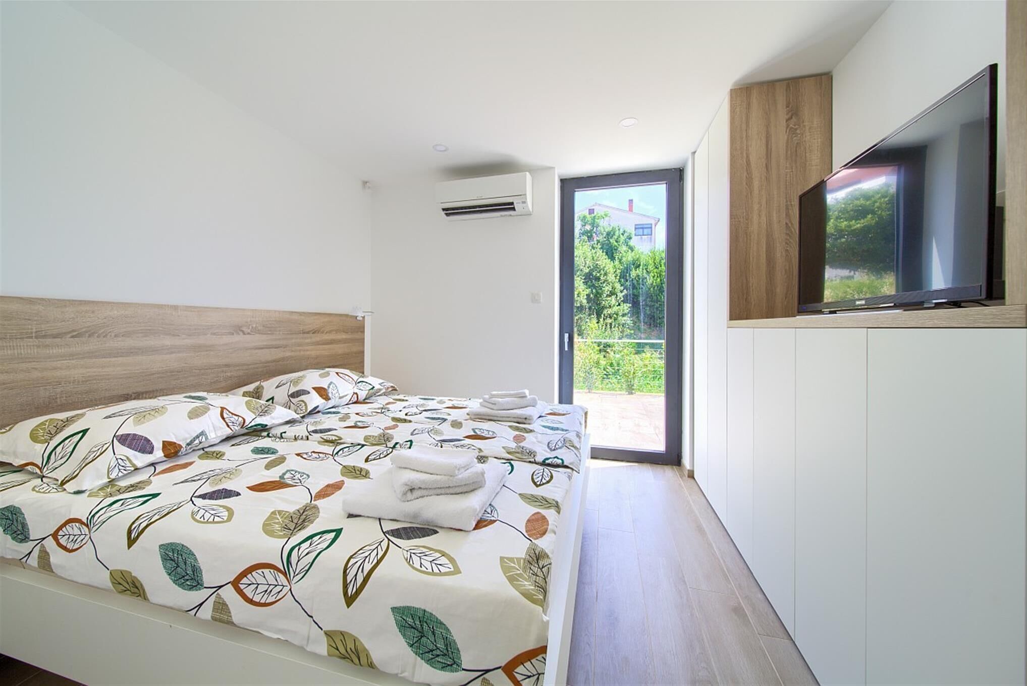 A modern bedroom with a large bed, a television mounted on the wall, and a sliding glass door leading to a balcony.