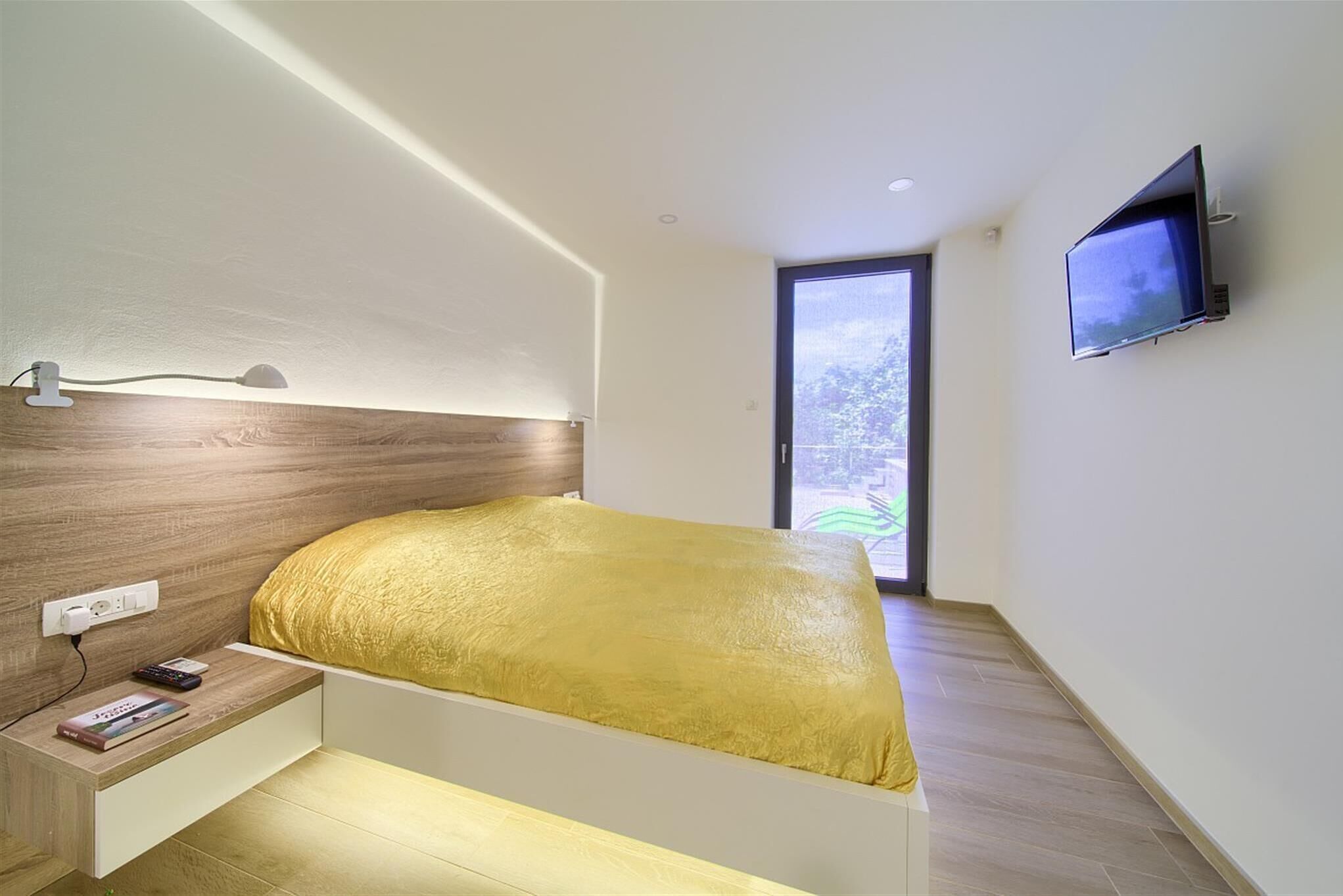 A modern bedroom with a bed, bedside table, TV, and a window.