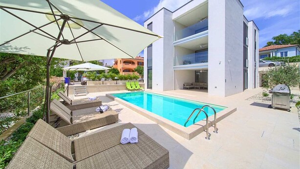 A modern pool area with a clear blue pool, sun loungers, and a large umbrella.