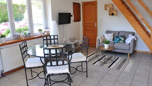 Un salón acogedor con un sofá gris, una mesa de comedor de cristal con sillas, una escalera de madera y vistas al jardín a través de ventanas grandes.
