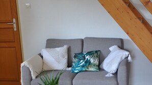 Un sofá gris con varios cojines, una pequeña mesa de madera con una planta y una alfombra a rayas sobre piso de baldosas.