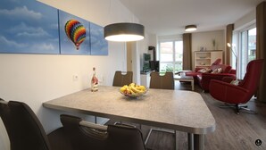 Uma sala de estar moderna com área de jantar, apresentando uma mesa grande com uma tigela de frutas, cadeiras vermelhas e uma televisão.