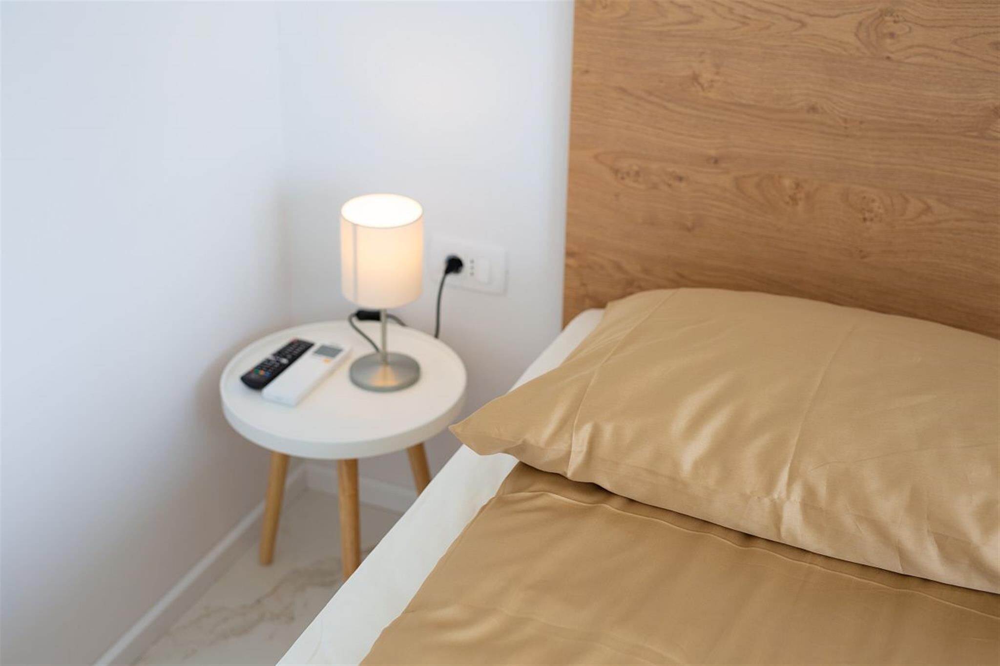A neatly made bed with a wooden headboard, a round bedside table with a lamp, and a remote control on the table.