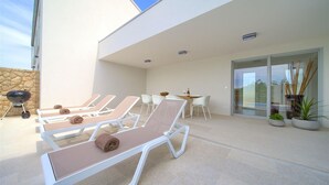 A modern outdoor lounge area with sun loungers, a barbecue grill, and a dining table with chairs.
