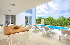 Een modern terras buiten met een houten tafel, witte stoelen en een zwembad.