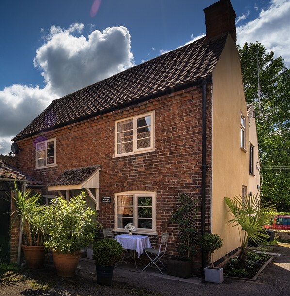 A brick house with a tiled roof, a small garden with potted plants, and a table with a chair outside.