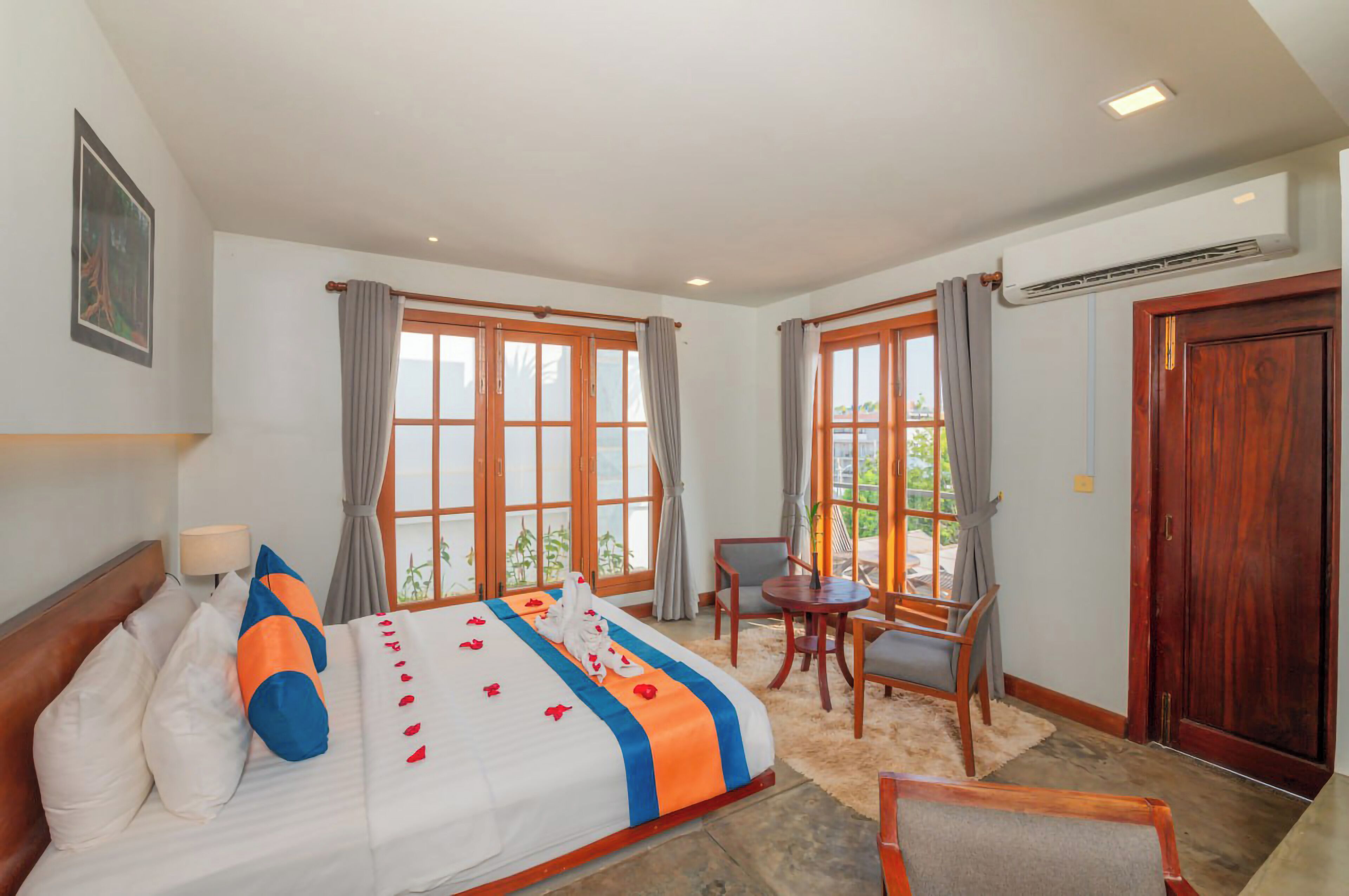 A hotel room with a bed, a desk, and a chair. There are windows with curtains, an air conditioning unit, and a wooden door.