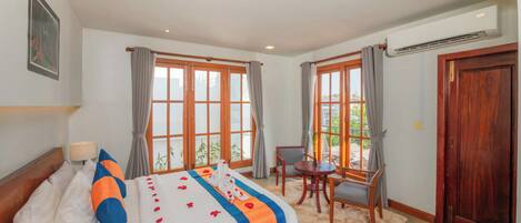 A hotel room with a bed, a desk, and a chair. There are windows with curtains, an air conditioning unit, and a wooden door.