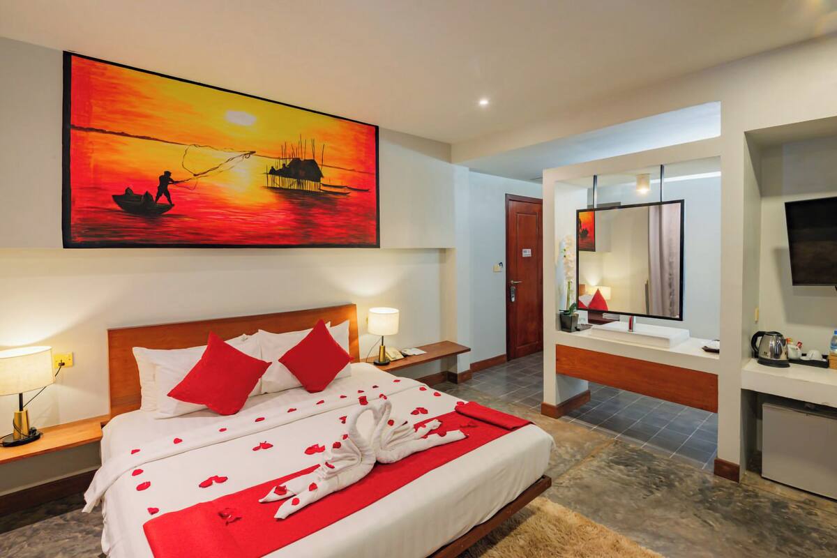 A hotel room with a bed, a painting of a sunset over water, a TV, and a small kitchenette.