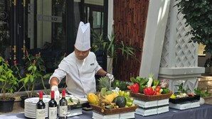 Un chef prépare des plats en plein air, sur une table où sont disposées des bouteilles de vin, des fruits et des légumes.