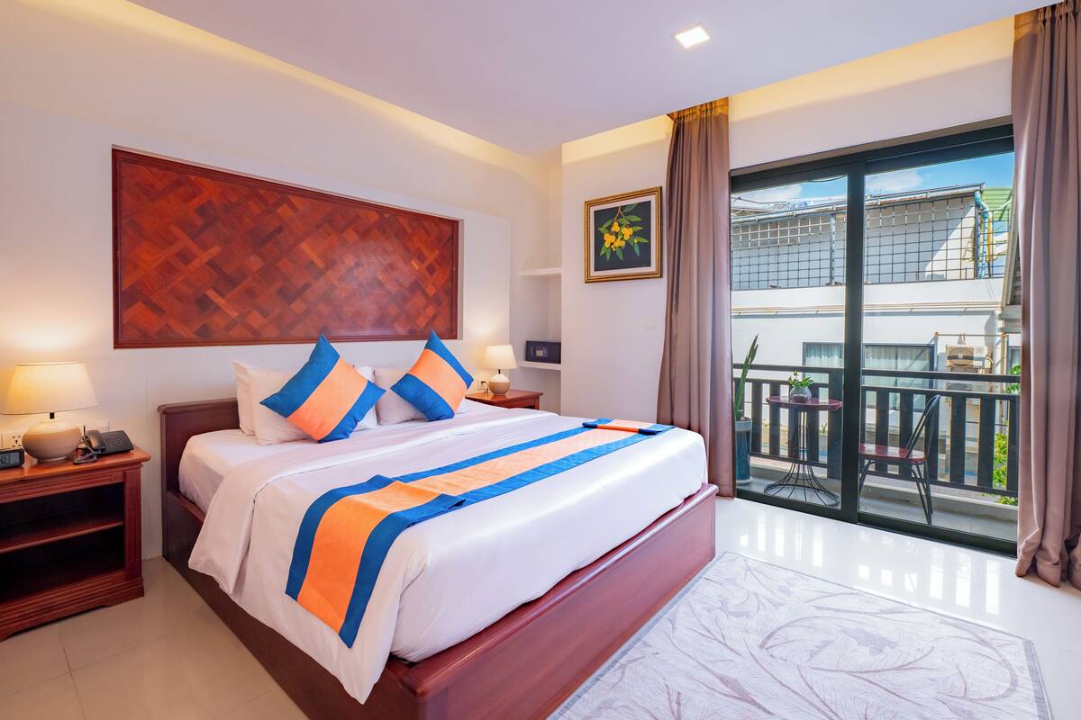A hotel room with a large bed, a nightstand with a lamp, a wall-mounted artwork, a balcony with a view, and a potted plant.