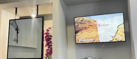 A modern bathroom with a large mirror, a potted orchid, and a flat-screen TV displaying a map.