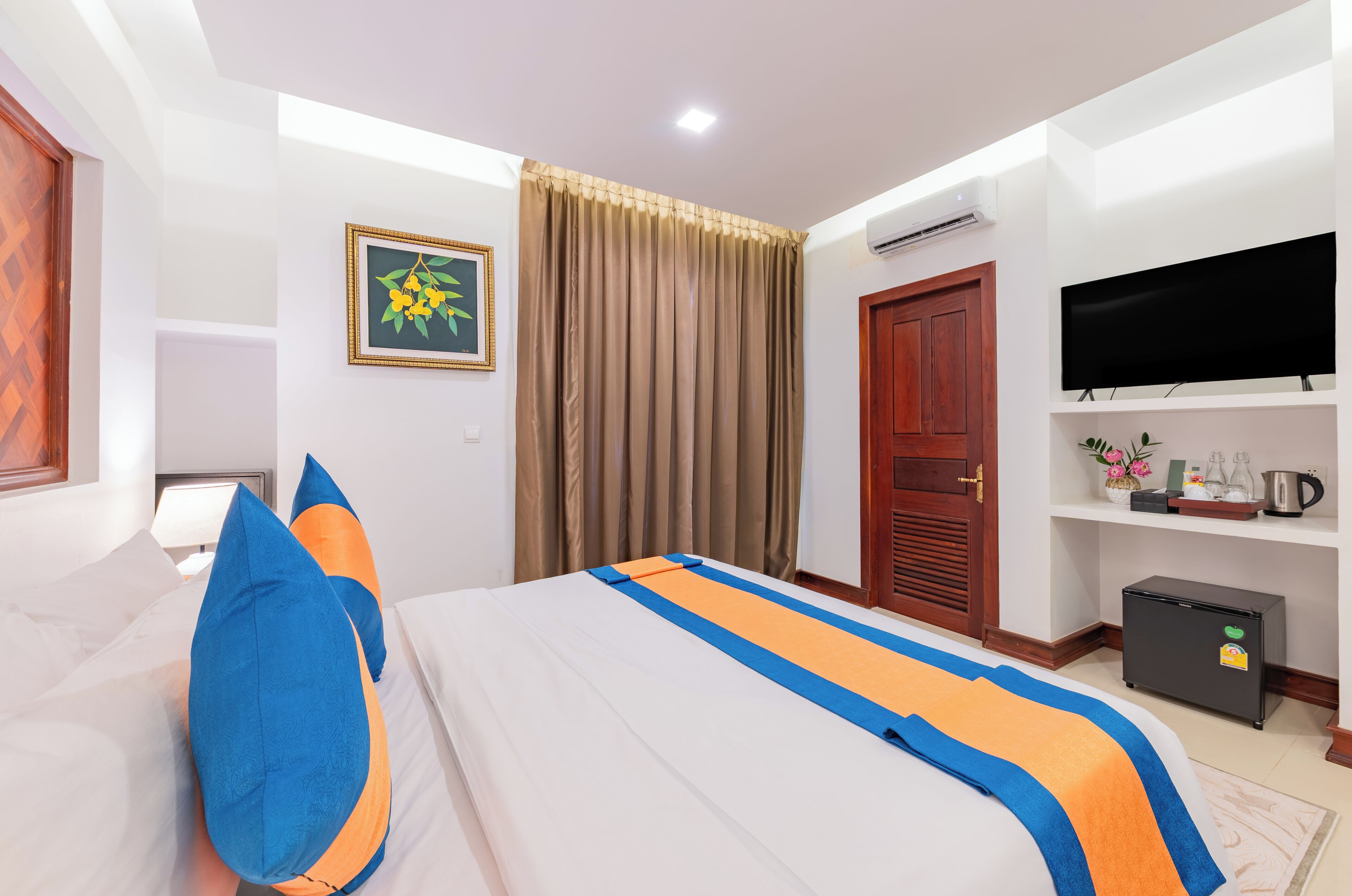 A hotel room with a bed, a TV, a wardrobe, a shelf with decorative items, and a small refrigerator.