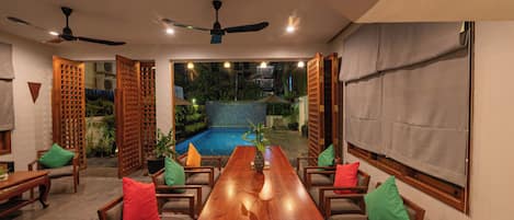 A dining area with a long wooden table, chairs with colorful cushions, a ceiling fan, and a view of a swimming pool and greenery.