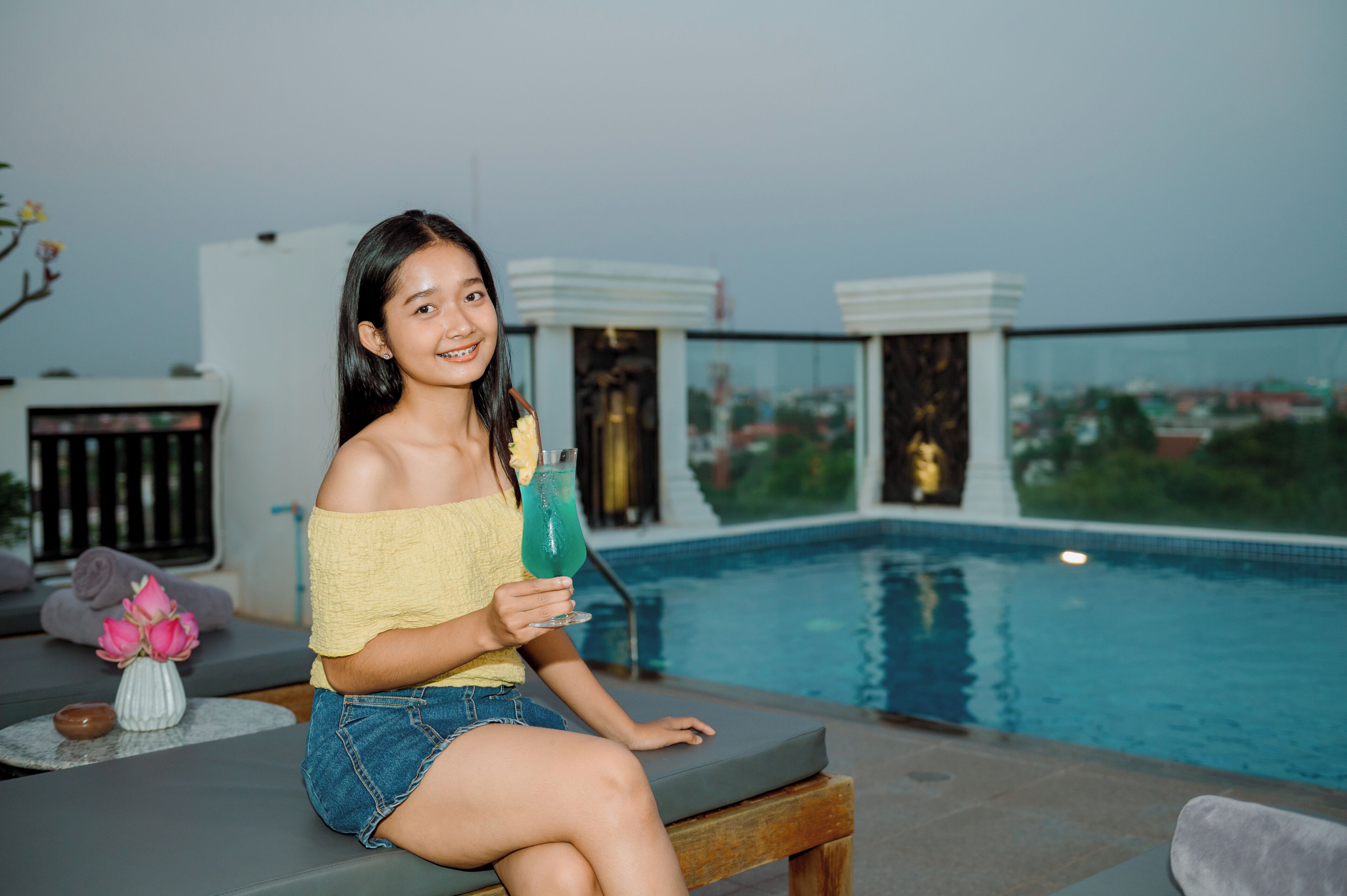 A person sitting by a pool with a drink, a sofa, and a vase of flowers.