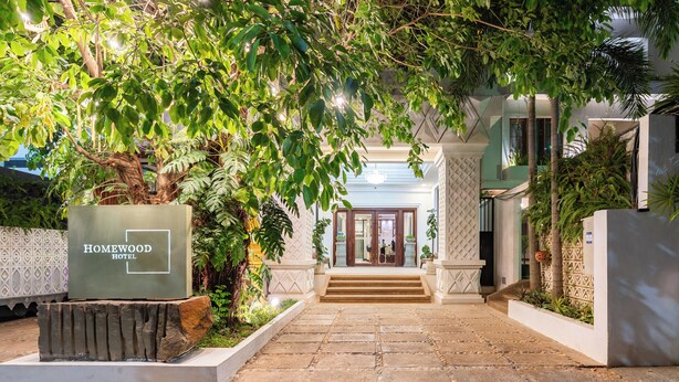 L’entrée d’un hôtel, avec un chemin de pierre menant aux portes, est bordée de verdure et un panneau indique : « HOMEWOOD HOTEL ».